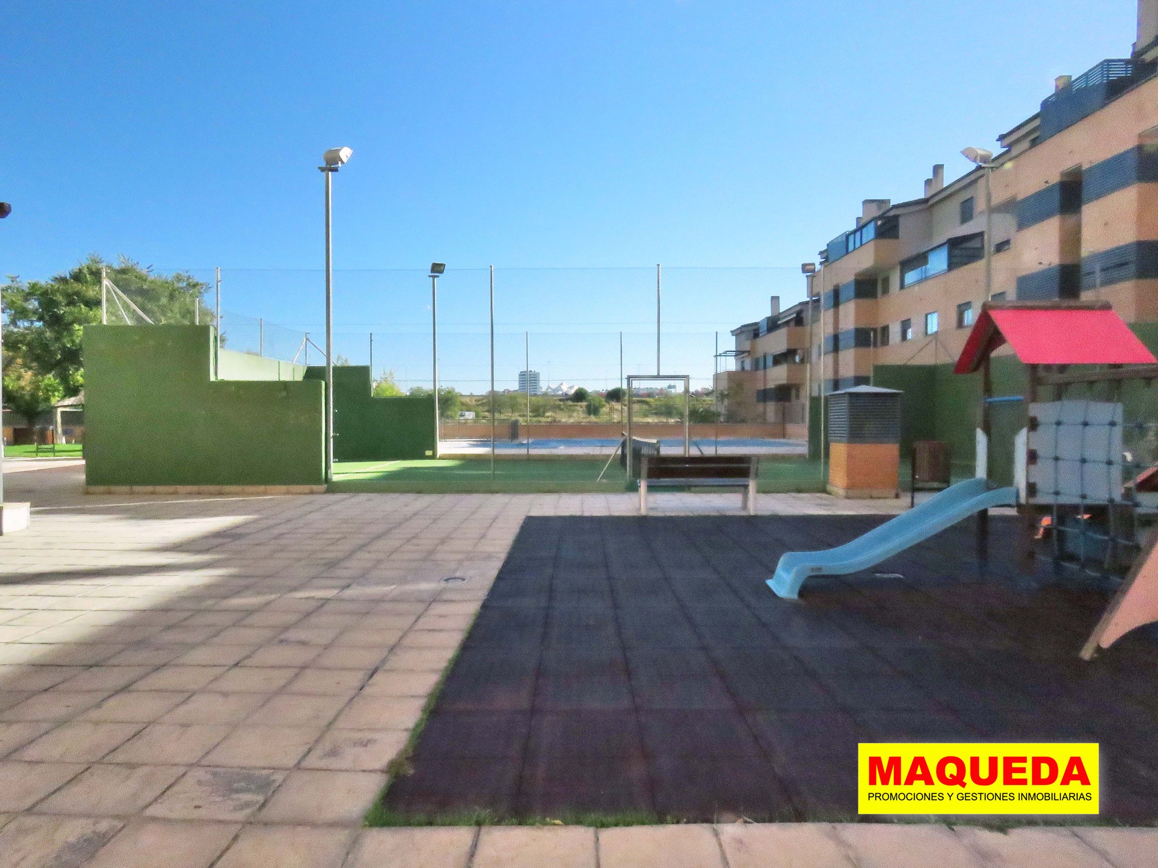 Interior de la urbanización con zona infantil y columpios en primer plano. Al fondo pista de pádel