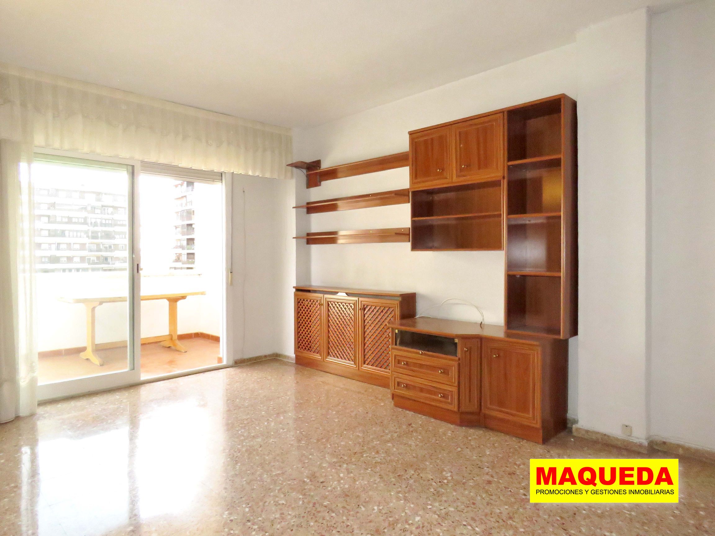 Mueble con estanterías en el salón comedor. A la izquierda puerta de acceso a la terraza con mesa. Al fondo edificios